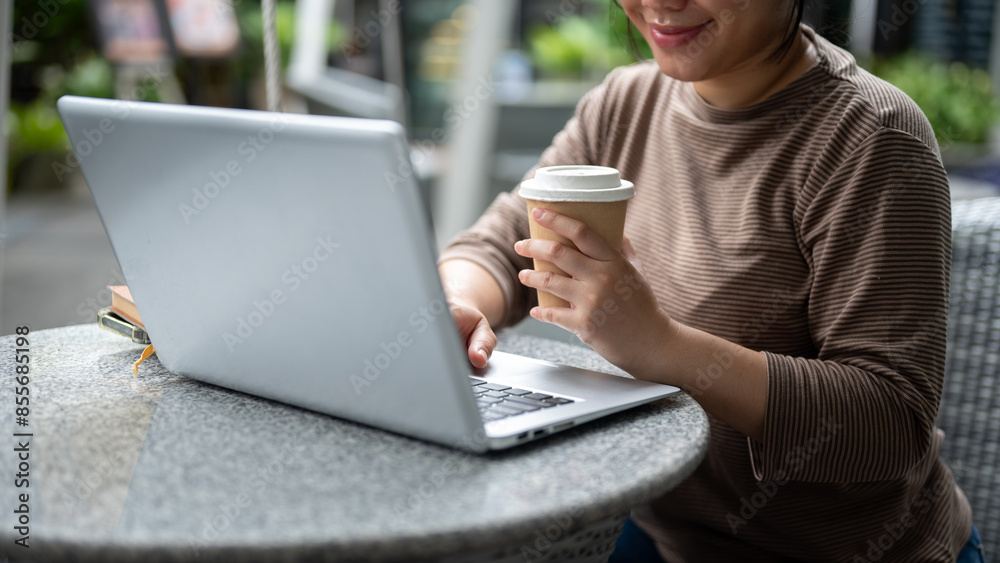 A cropped image of an Asian woman having coffee while working remotely on her laptop at a cafe.