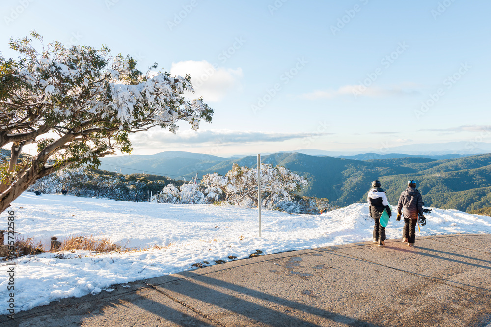 Fototapeta premium Mount Buller in Victoria, Australia. A pair of snowboarders walk across the snow to admire the view of the mountains.