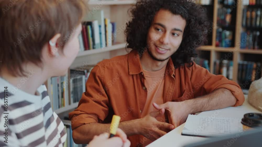Young middle-eastern student with curly hair sitting with copybook at desk in the library and chatting with female group mate about college life. Over the shoulder view Young middle-eastern student with curly hair sitting with copybook at desk in the library and chatting with female group mate about college life. Over the shoulder view