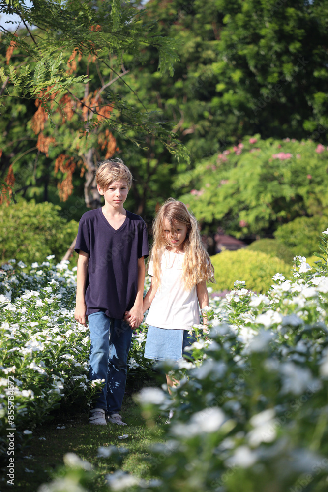 Fototapeta premium Happy family, brother and sister hold hands, walk through beautiful flower garden together. Joyful kids, boy and girl playing outdoor together at public park. Children enjoy spending time outside.