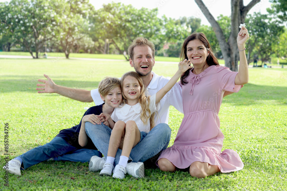 Happy family sitting on green grass and waving hands at park, smiling father, mother, son and daughter spending time together outdoors in beautiful garden on holiday. Parents and kids have fun outside Happy family sitting on green grass and waving hands at park, smiling father, mother, son and daughter spending time together outdoors in beautiful garden on holiday. Parents and kids have fun outside