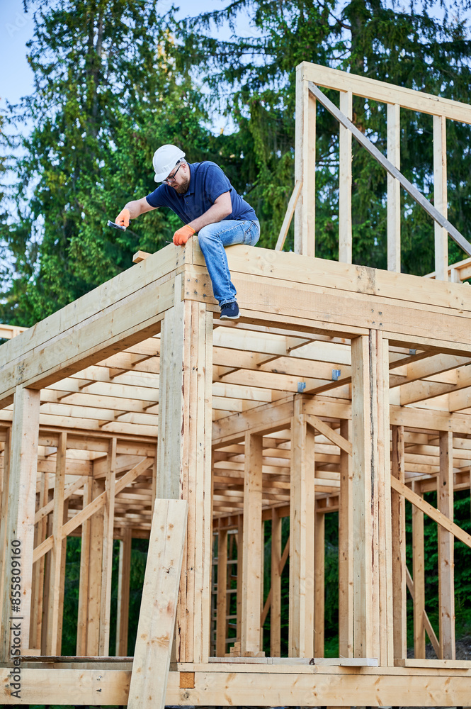 Carpenter building wooden, two-story frame house near woods. Bearded man with spectacles hammering nail while wearing protective helmet. The concept of contemporary ecological construction. Carpenter building wooden, two-story frame house near woods. Bearded man with spectacles hammering nail while wearing protective helmet. The concept of contemporary ecological construction.