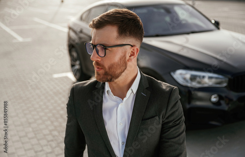 Handsome smart guy in glasses. Businessman in suit is near his black car outdoors © standret