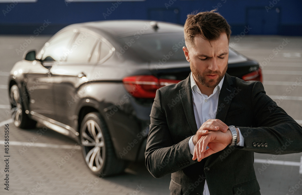 Checks the time. Businessman in suit is near his black car outdoors Checks the time. Businessman in suit is near his black car outdoors