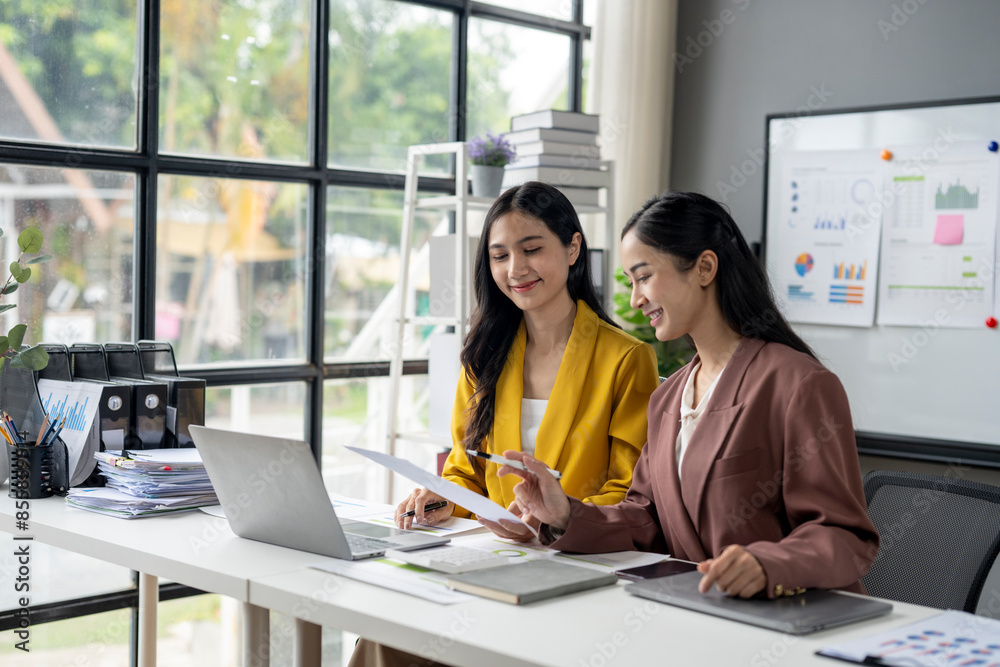 Two women are sitting at a desk with a laptop and papers in front of them Two women are sitting at a desk with a laptop and papers in front of them