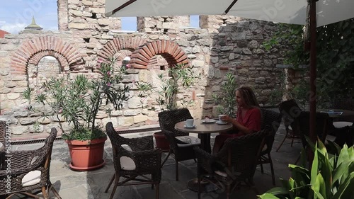  Woman sitting in a cafe at a table on the summer terrace, drinking coffee, eating cake, near the ruins of an old church in the old town of Nessebar, Bulgaria in summer. photo