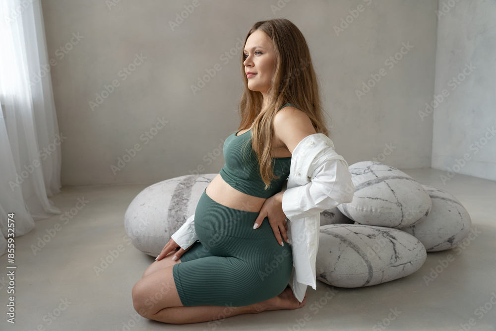 Sporty beautiful pregnant healthy woman with blonde hair wearing green sportswear and white shirt sitting indoors on floor in front of gray pillows looking away in anticipation of happy motherhood Sporty beautiful pregnant healthy woman with blonde hair wearing green sportswear and white shirt sitting indoors on floor in front of gray pillows looking away in anticipation of happy motherhood