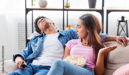 A girl sits on a couch with a bowl of popcorn in her lap, looking annoyed as the guy beside her sleeps while watching TV. He is holding a remote control in his hand.