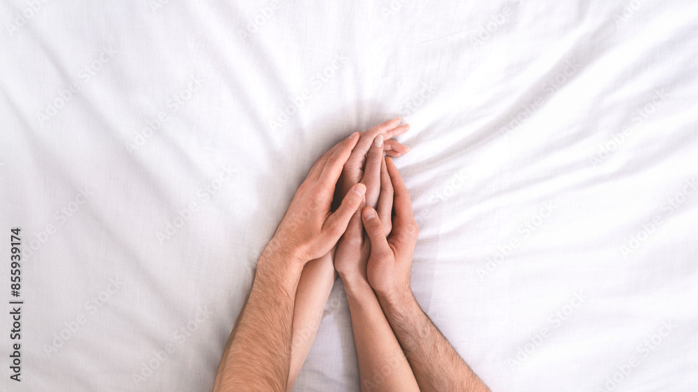 An overhead close-up shot of two hands, one male and one female, intertwined on white bedding. The hands are positioned in a loving and intimate way, suggesting a romantic or intimate connection An overhead close-up shot of two hands, one male and one female, intertwined on white bedding. The hands are positioned in a loving and intimate way, suggesting a romantic or intimate connection