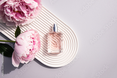A beautiful bottle of spray or perfume lies on a white tray in the form of an arch relief with delicate pink peonies. top view. A blank bottle mockup.