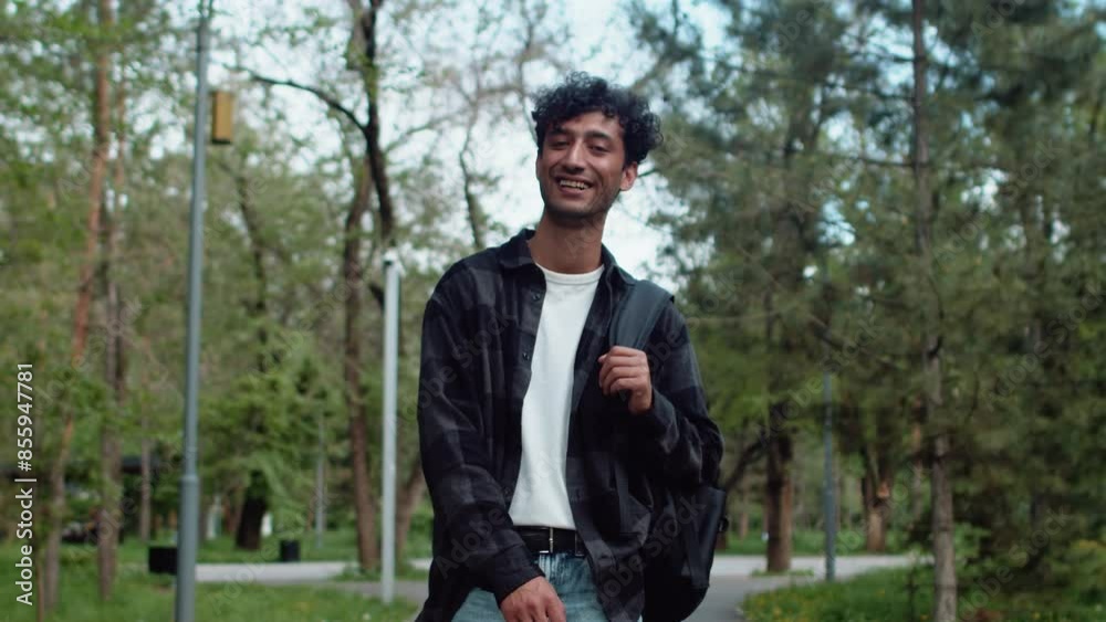 Young curly man in casual clothes with a backpack walking in an alley of the city park, then turning around and smiling. Concept of carefree urban life Young curly man in casual clothes with a backpack walking in an alley of the city park, then turning around and smiling. Concept of carefree urban life