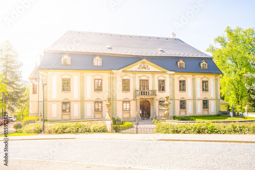 Photo of Potstejn, a charming baroque chateau in Czechia. Yellow facade, dark blue roof, surrounded by lush greenery, trees, and a garden.