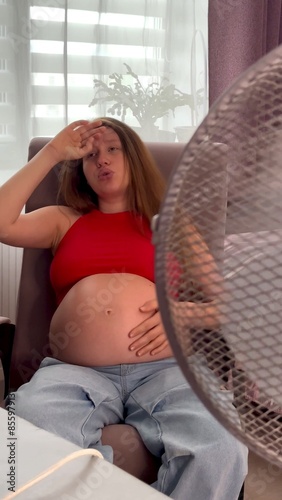 Young pregnant woman using electric fan at home in living room, sitting on couch cooling off during hot weather, suffering from heat, high temperature