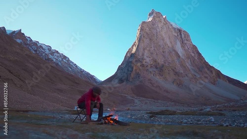 Indian male tourist fixing the woods of the bonfire in front of the mountain in Zanskar Valley at Ladakh, India. Tourist warming himself with fire in cold Himalayas in India.	 photo