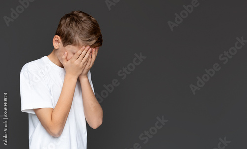 The young boy in a white tee covers his eyes with hands, expressing fear, shame, or a desire not to see, isolated on a grey background
