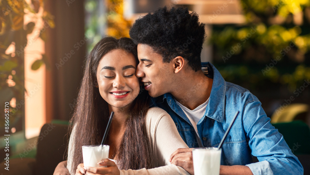 African American young guy and girl are sharing a tender moment as they kiss at a table in a bustling coffee shop. The couple is engrossed in each other, surrounded by coffee cups and a cozy ambiance. African American young guy and girl are sharing a tender moment as they kiss at a table in a bustling coffee shop. The couple is engrossed in each other, surrounded by coffee cups and a cozy ambiance.
