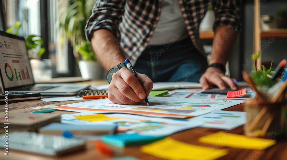 Close-up of a person working on a project at a cluttered desk with charts and graphs, illustrating a busy and productive workspace. Close-up of a person working on a project at a cluttered desk with charts and graphs, illustrating a busy and productive workspace.