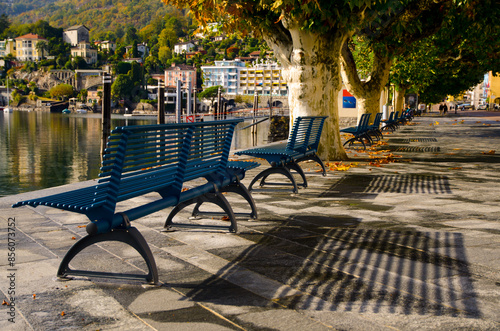 Empty Benches with Shadows on the Waterfront to Lake Maggiore in Autumn with Trees and Mountain View in a Sunny Day in City of Ascona, Ticino, Switzerland.