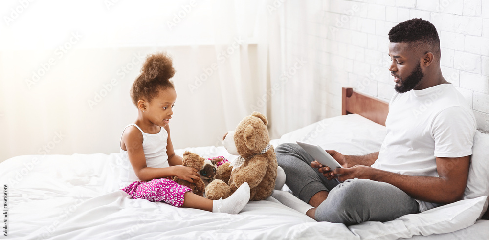 Black father sits on a white bed while his daughter plays with a teddy bear. He is looking down at a tablet in his lap, while she is looking at him. The bed is white and the room is bright. Black father sits on a white bed while his daughter plays with a teddy bear. He is looking down at a tablet in his lap, while she is looking at him. The bed is white and the room is bright.