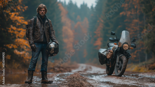 A motorcyclist standing on a dirt road, with a motorcycle parked beside him. The motorcyclist is wearing a leather jacket, jeans, and boots, and is holding a helmet in his hand