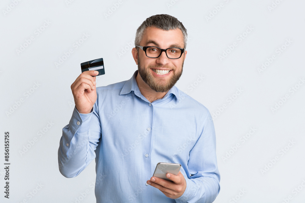 A man is depicted holding a credit card in one hand and a cell phone in the other hand. He appears to be engaged in a transaction or making a purchase online. A man is depicted holding a credit card in one hand and a cell phone in the other hand. He appears to be engaged in a transaction or making a purchase online.