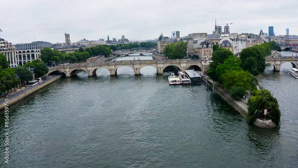 Fototapeta premium Aerial view of Paris featuring the Seine River and several bridges, including the Pont Neuf. The city showcases its historic architecture, with green trees by the river and boats moored at the docks.