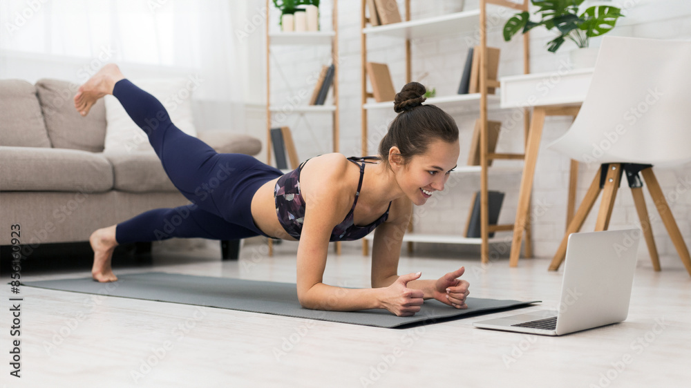 A woman in athletic wear is doing a plank exercise on a yoga mat while watching a laptop. She is in her living room with a couch and bookshelf in the background. A woman in athletic wear is doing a plank exercise on a yoga mat while watching a laptop. She is in her living room with a couch and bookshelf in the background.