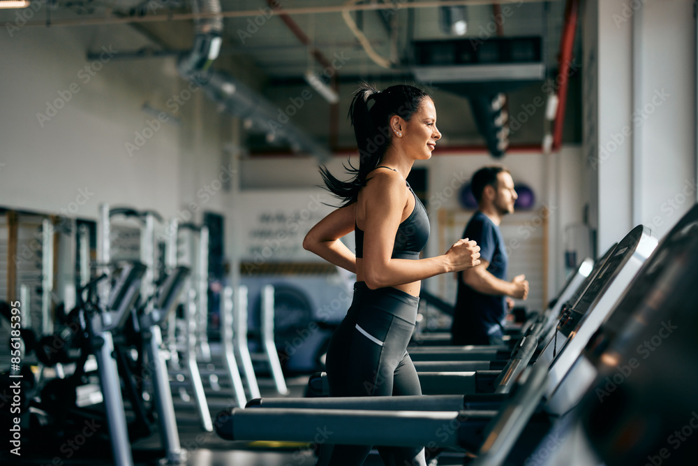 Side profile of a female running on a treadmill at the gym, a male running in the background. Side profile of a female running on a treadmill at the gym, a male running in the background.