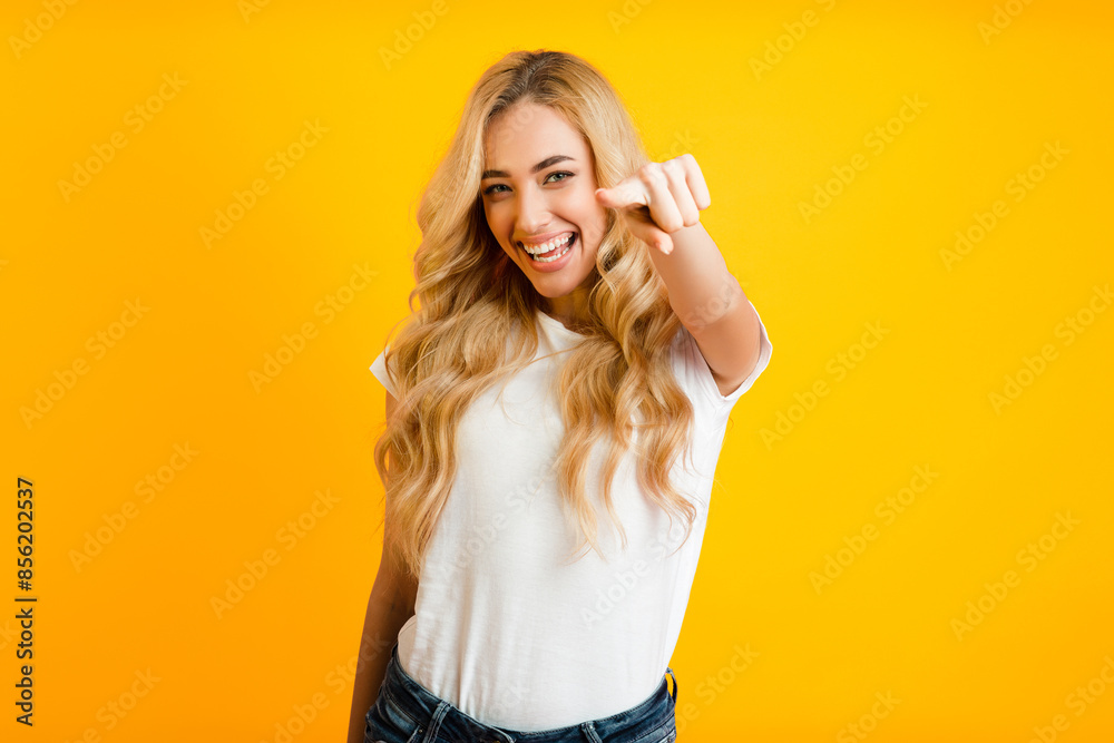 A young woman with long, blonde hair is smiling and pointing directly at the camera. She is wearing a white t-shirt and jeans and is standing in front of a solid yellow background. A young woman with long, blonde hair is smiling and pointing directly at the camera. She is wearing a white t-shirt and jeans and is standing in front of a solid yellow background.
