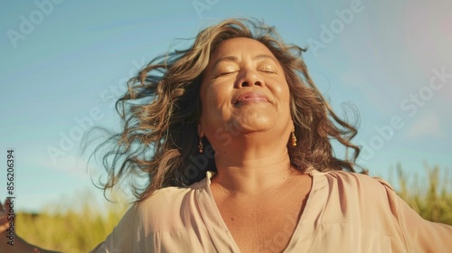 Woman with closed eyes smiling in a field with wind-blown hair wearing a light-colored blouse and earrings with a blue sky and green grass in the background.