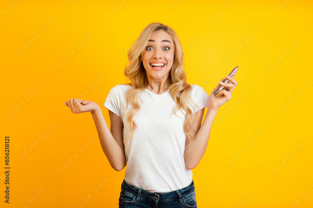 A blonde woman stands in front of a bright yellow background with a surprised expression on her face, holding a smartphone in one hand, and her other hand outstretched in a gesture of disbelief A blonde woman stands in front of a bright yellow background with a surprised expression on her face, holding a smartphone in one hand, and her other hand outstretched in a gesture of disbelief