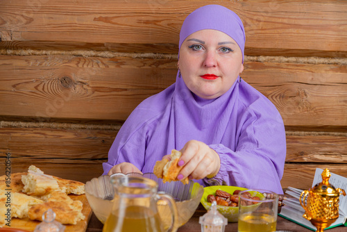 A Muslim woman in a lilac hijab at the table eats lamb with flatbread on the holiday of breaking the fast of Eid al-Fitr