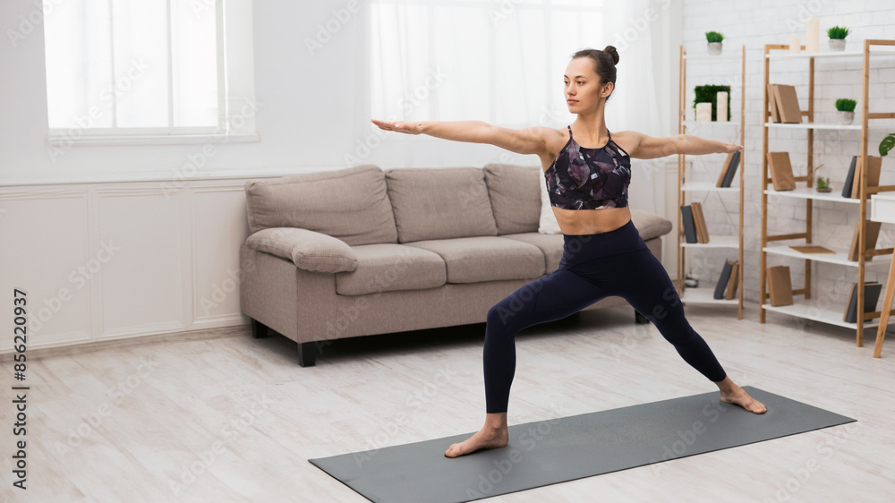 Obraz premium A woman is practicing yoga in her living room, stretching her arms and legs while standing on a yoga mat.