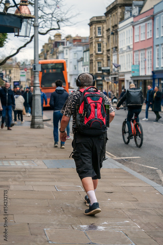 Pedestrian with a red backpack and headphones walking on a busy city street surrounded by cyclists and other people, urban life daily, city exploration, mobility lifestyle and travel