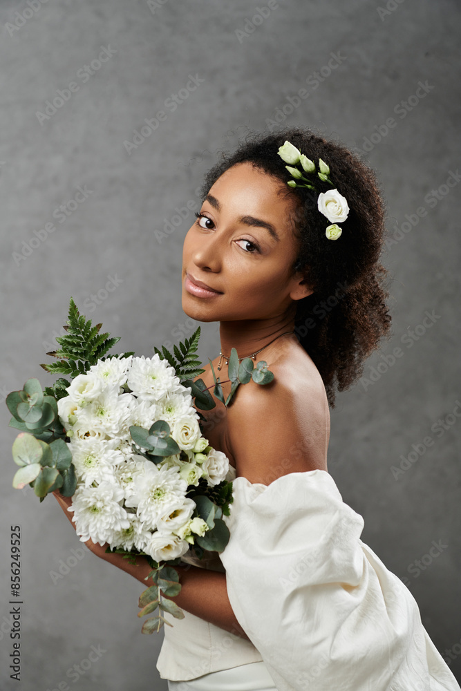 African American bride in white dress with flowers in her hair holds bouquet against grey background.