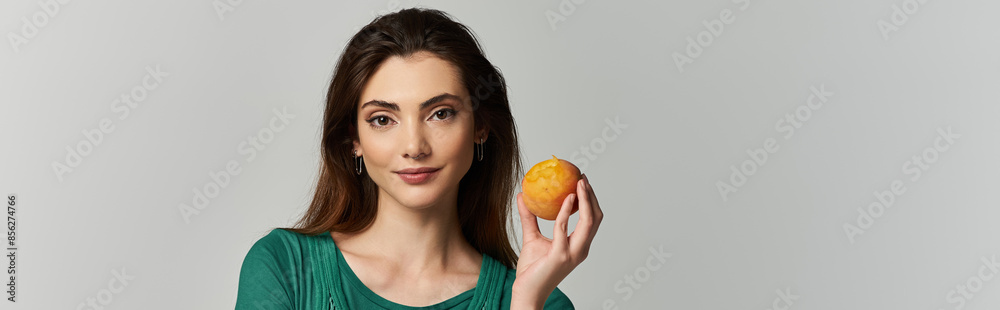 A young woman in an emerald green top holds a peach and smiles at the camera. A young woman in an emerald green top holds a peach and smiles at the camera.