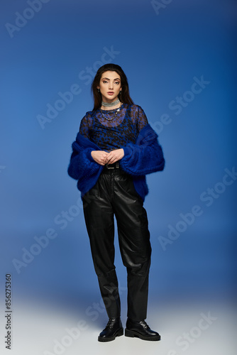 A young woman in black leather trousers and a blue fuzzy cardigan stands against a blue backdrop.