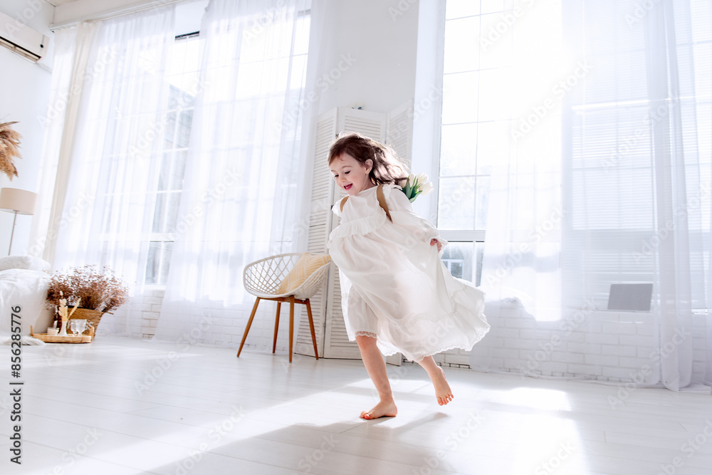 Happy little girl in flowing white dress runs barefoot in bright, sunlit room. Airy space, interior decorated in minimalist style, enhances feeling of freedom, joy. Happiness childhood. Boho style Happy little girl in flowing white dress runs barefoot in bright, sunlit room. Airy space, interior decorated in minimalist style, enhances feeling of freedom, joy. Happiness childhood. Boho style