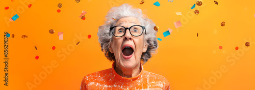 A surprised elderly woman with gray hair and glasses, wearing an orange shirt, stands against a vibrant orange background with confetti falling around her.