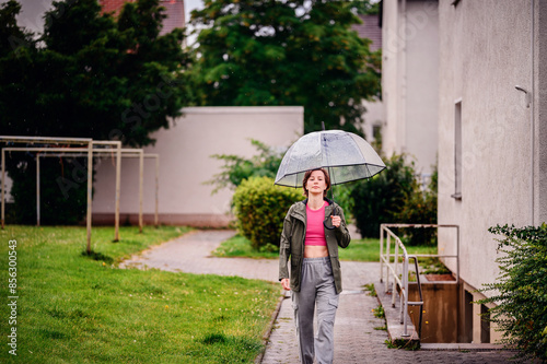 Young woman wearing a pink top and green jacket, walking outdoors with a transparent umbrella on a rainy day. The scene captures a moment of serenity and calm.