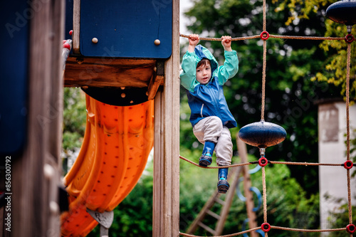 Young child wearing a teal and blue raincoat, climbing on a playground climbing frame on a rainy day. The scene captures the spirit of adventure and play.