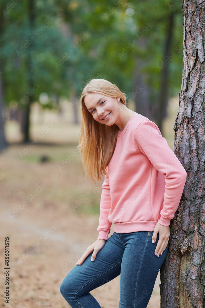 Blue-eyed blonde in a pink hoodie walks in a pine forest. Portrait of a joyful young woman enjoying in autumn park.