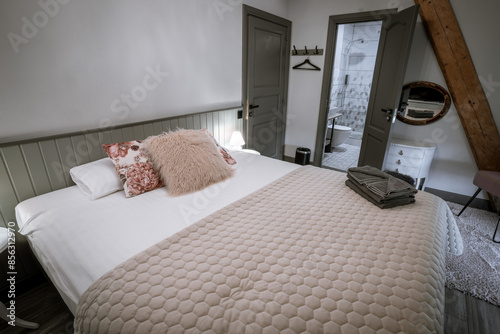 Blome, Latvia - September 11, 2023 - A cozy bedroom with a neatly made bed, floral and furry pillows, folded towels, bedside tables with lamps, and an open door leading to a modern bathroom. photo