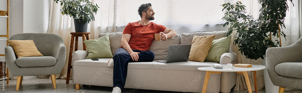 A man with a beard sits on a sofa in a well-lit living room, working on his laptop while enjoying a cup of coffee. A man with a beard sits on a sofa in a well-lit living room, working on his laptop while enjoying a cup of coffee.