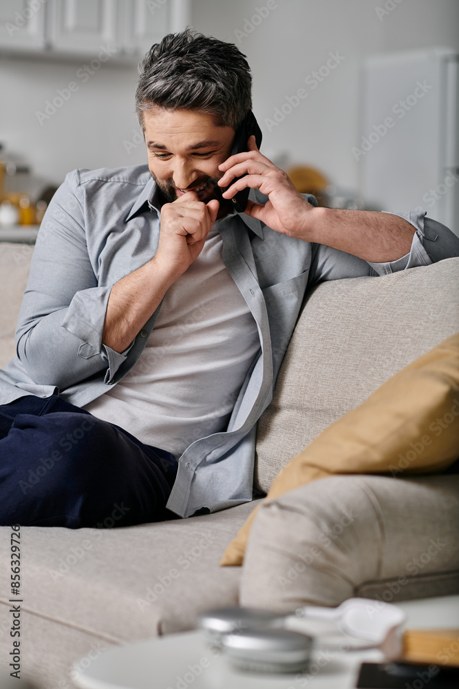 A bearded man in casual attire sits on a couch and talks on the phone while smiling. He appears to be working remotely from home. A bearded man in casual attire sits on a couch and talks on the phone while smiling. He appears to be working remotely from home.