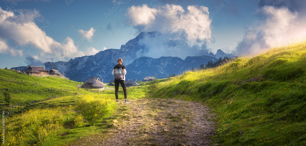 Girl on the rural road with yellow flowers and green grass in beautiful alpine mountain valley at sunset in summer. Young woman in old alpine village, sky with clouds. Travel and Hiking. Slovenia Girl on the rural road with yellow flowers and green grass in beautiful alpine mountain valley at sunset in summer. Young woman in old alpine village, sky with clouds. Travel and Hiking. Slovenia
