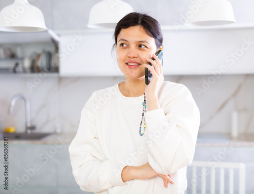 Calm and cheerful young woman with dark hair dressed in comfortable white clothing, talking amiably on mobile phone while standing in cozy home kitchen © JackF