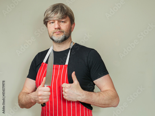 Male butcher in red and white classic apron and black shirt holding knife. Light color background. Man in his 40s with grey beard and hair. He is smiling and looking at the camera. Meat industry.