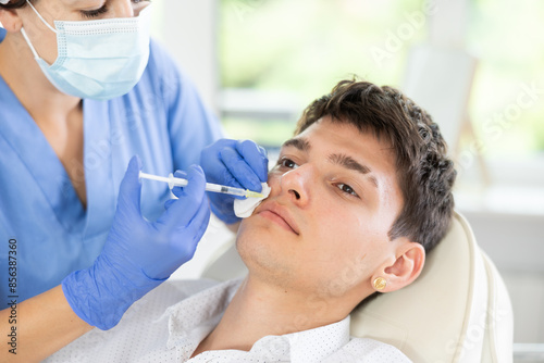 Female doctor conducting face revitalization procedure for young man patient by micro-injection method