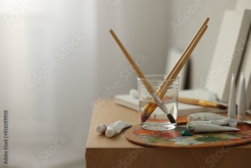 Artist's palette, brushes in glass of water and paints on wooden table indoors. Space for text